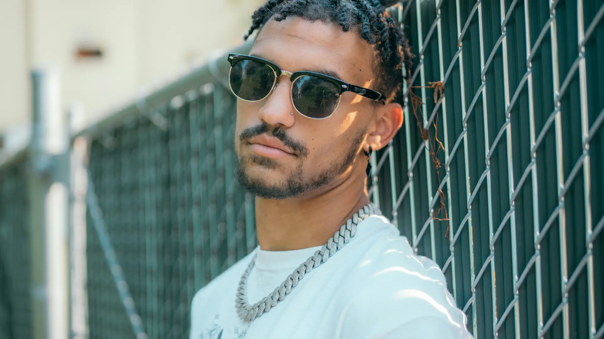 Homem negro vestindo uma camiseta branca, óculos de sol e uma corrente de prata posa para foto encostado em uma grade