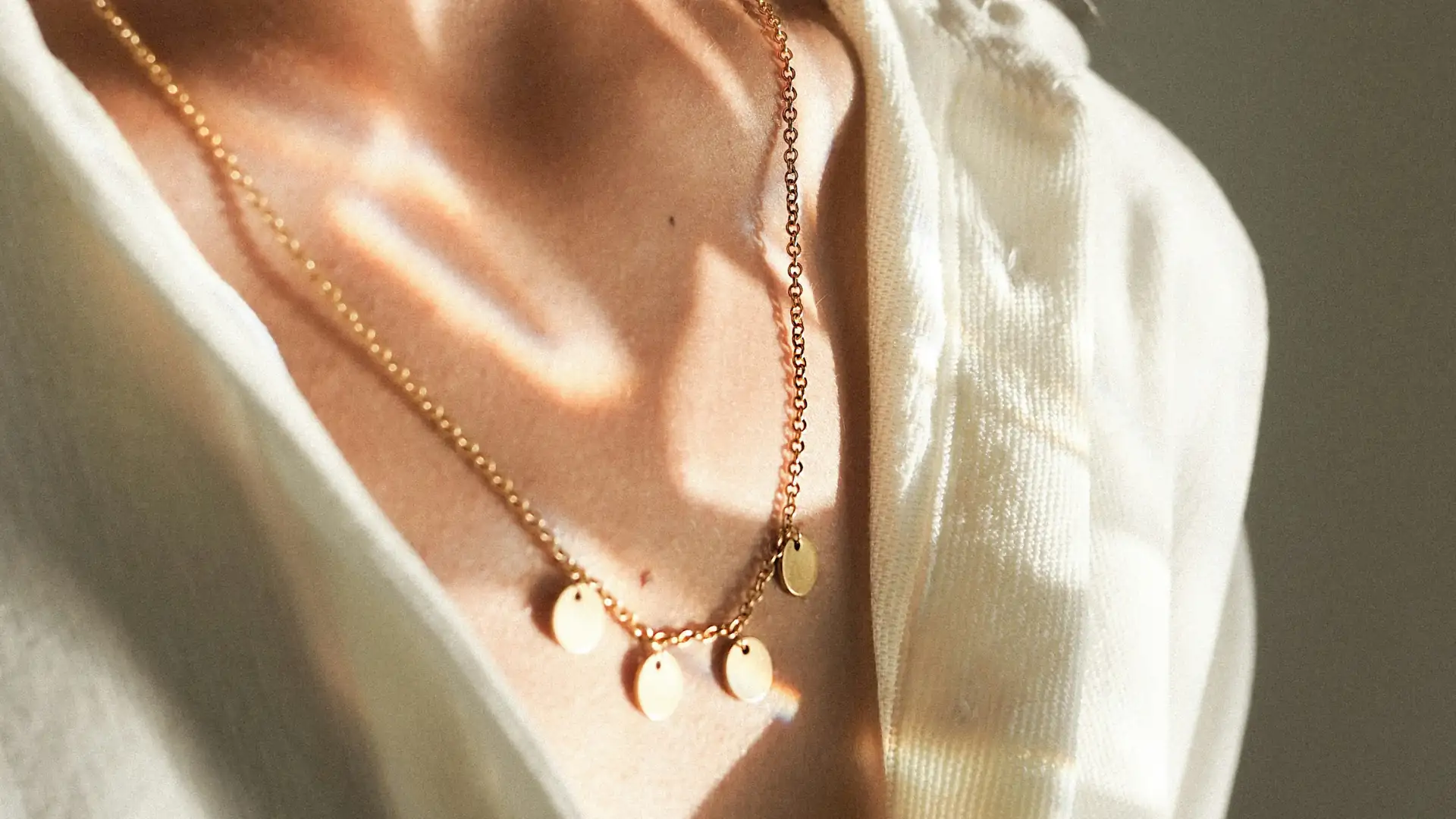 Colar dourado delicado com pequenos pingentes circulares refletindo a luz, sobre a pele de uma mulher branca que veste uma camisa clara e leve.