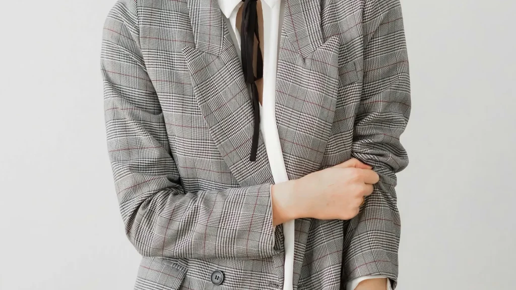 Close de blazer cinza com estampa xadrez clássica, sobreposto a uma camisa branca com detalhe de laço fino preto no pescoço.