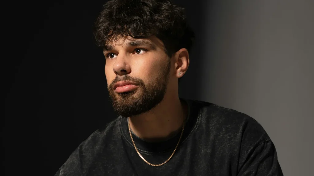 Homem jovem de barba e cabelo cacheado usando uma fina corrente de ouro masculina sobre camiseta escura.