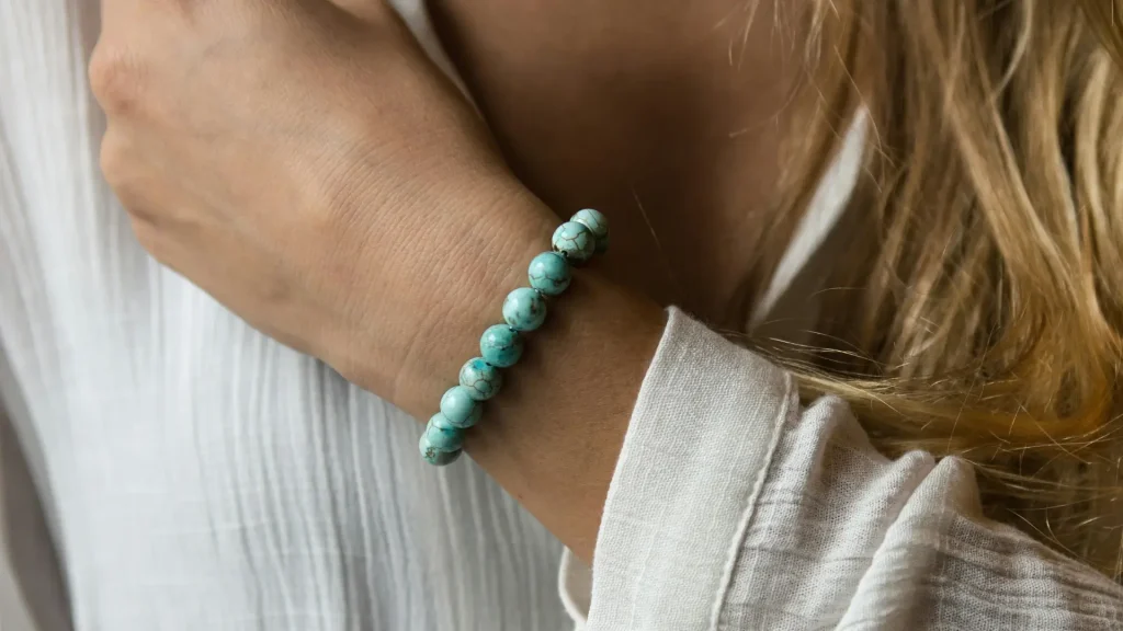 Pulso feminino adornado com uma pulseira de contas redondas em pedra turquesa sobre o punho de uma blusa branca de tecido leve.