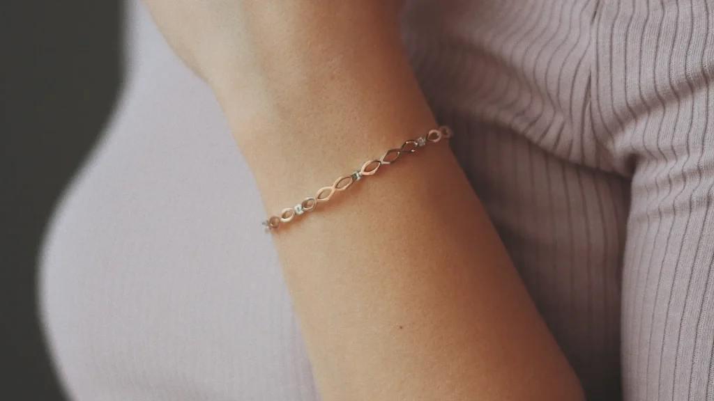 Detalhe de braço feminino usando uma pulseira fina de ouro rose com elos vazados em formato geométrico, combinando com blusa canelada.