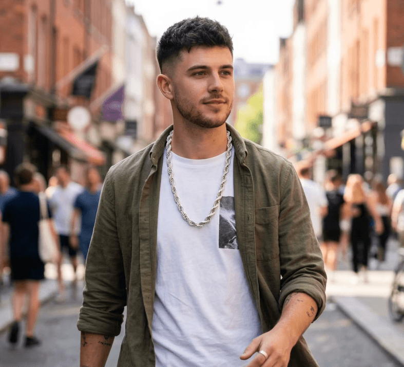 Jovem de camiseta branca e camisa verde aberta caminhando em uma rua de cidade. Ele usa um cordão baiano grosso de prata e anel, com tatuagens nos braços.