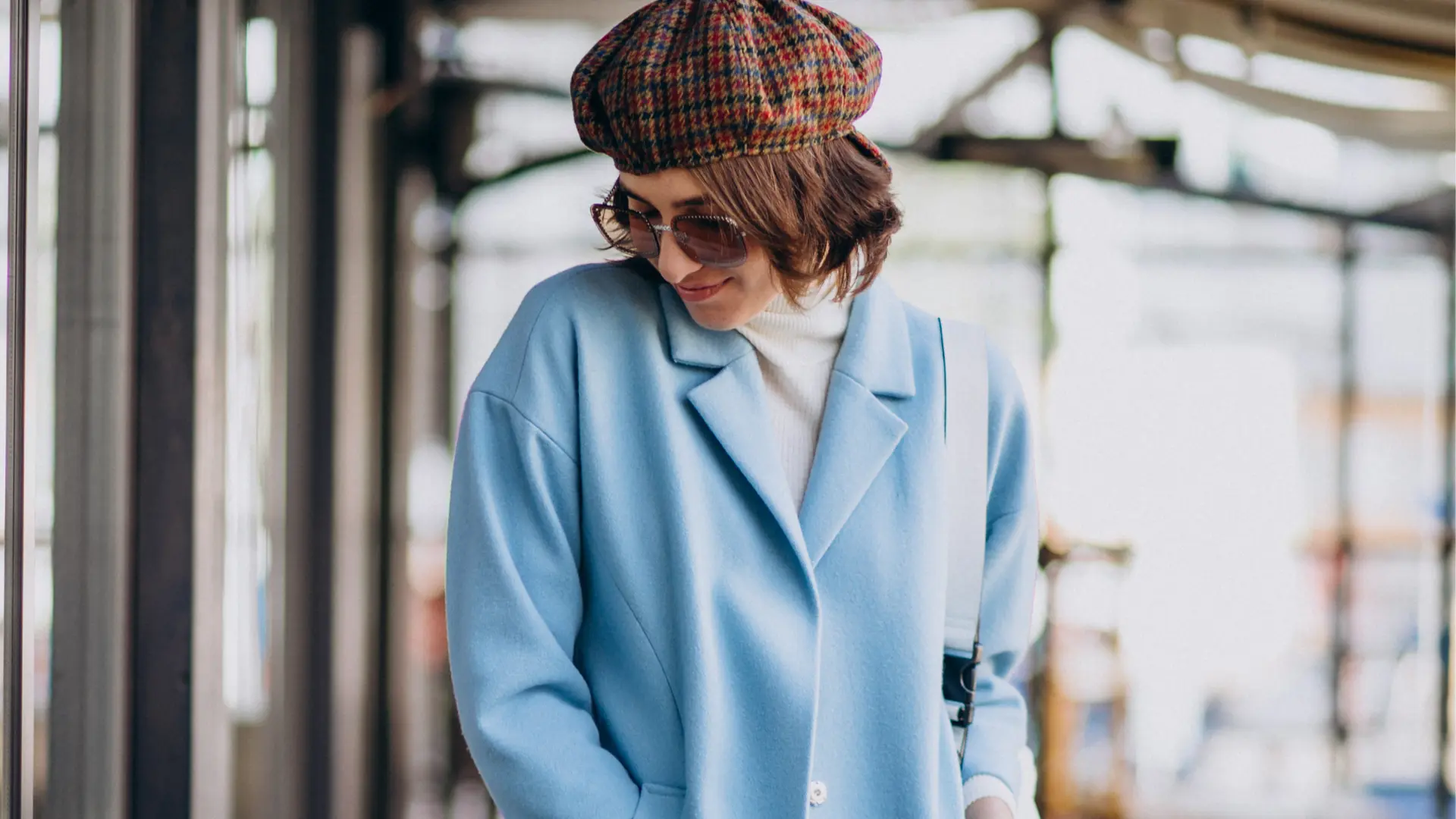 Mulher jovem sorridente usando casaco azul claro sobre blusa branca e boina xadrez, ilustrando cores que combinam com azul em looks de inverno.