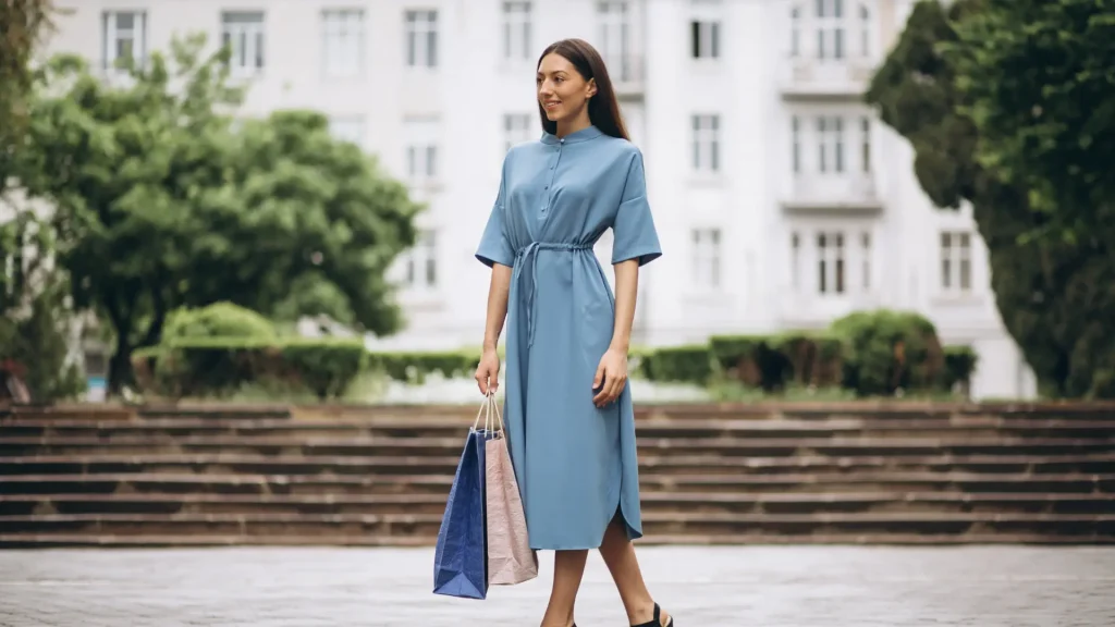 Mulher jovem com vestido midi azul jeans, de gola alta e amarração na cintura, segurando sacolas de compras. O look destaca cores que combinam com azul em um visual monocromático.