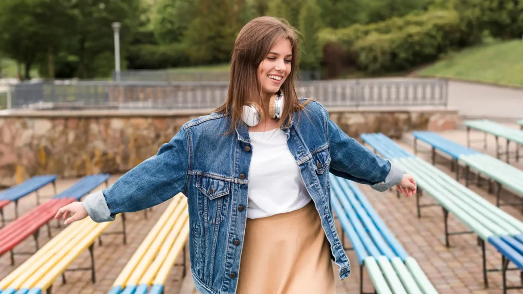 Mulher sorridente com jaqueta jeans azul sobre blusa branca e saia bege. O look casual e moderno exemplifica as cores que combinam com azul usando tons neutros e terrosos.