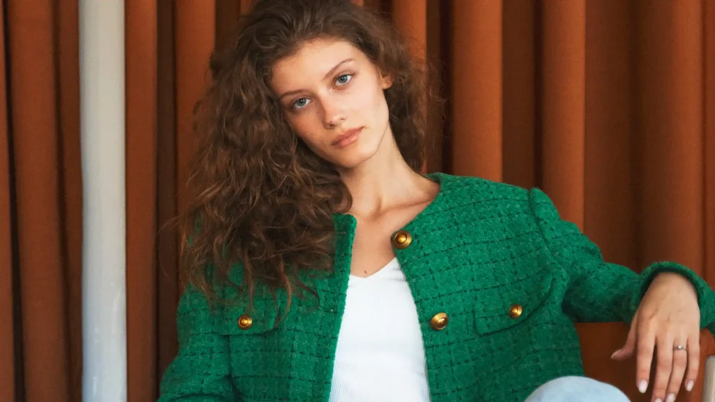 Mulher de cabelos ondulados vestindo um casaco de tweed verde vibrante sobre blusa branca, destacando o contraste de cores que combinam com verde.