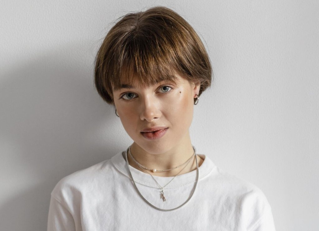 Close-up de uma mulher com cabelo castanho curto e olhos claros, olhando diretamente para a câmera contra uma parede branca. Ela veste uma camiseta branca de gola redonda. Em seu pescoço, ela exibe várias camadas de correntes de prata feminina, incluindo uma delicada com uma pequena pérola central e uma mais grossa com um pingente geométrico de prata. Ela também usa pequenos brincos de argola de prata.