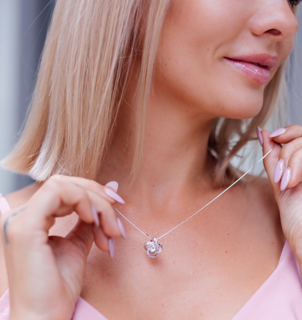 Uma mulher loira com unhas rosa pálido segurando uma delicada corrente de prata feminina de malha fina, ajustando-a no pescoço. O colar apresenta um pingente complexo em forma de nó cravejado de pequenas pedras de zircônia, com uma pedra maior no centro.