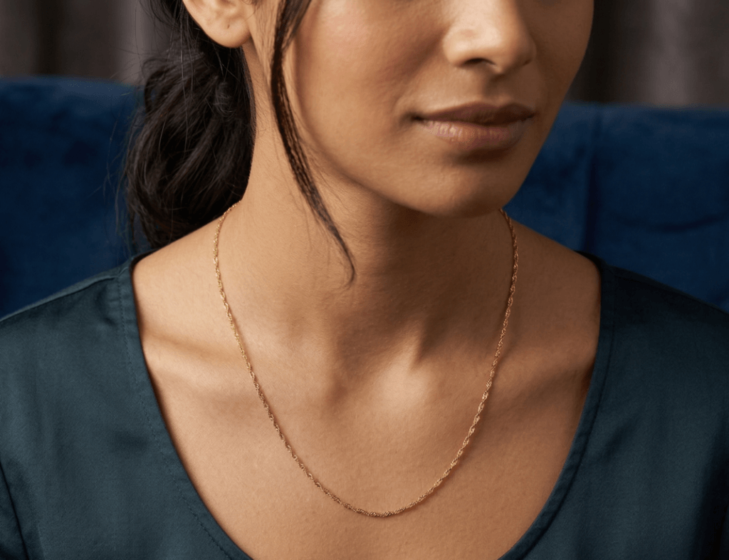 Close no pescoço de uma mulher vestindo uma blusa de seda verde-escura. Ela usa uma única corrente Singapura de ouro, que cai delicadamente sobre o colo. O fundo é de veludo azul-marinho.