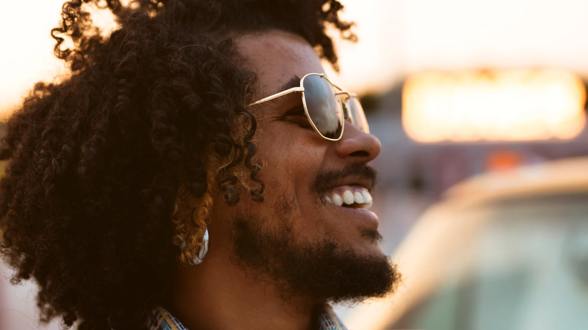Homem sorridente com cabelo crespo volumoso e óculos de sol, exibindo um dos estilos de barba curta e aparada, ideal para um visual moderno e despojado.