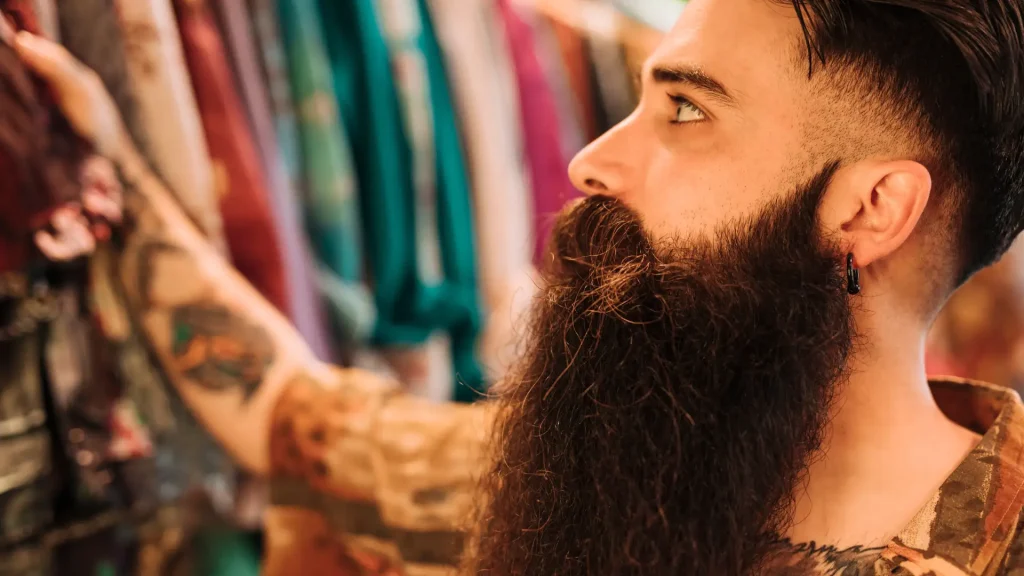Homem de perfil com barba longa e volumosa, representando estilos de barba lenhador (lumberjack) em um ambiente vintage com roupas ao fundo.