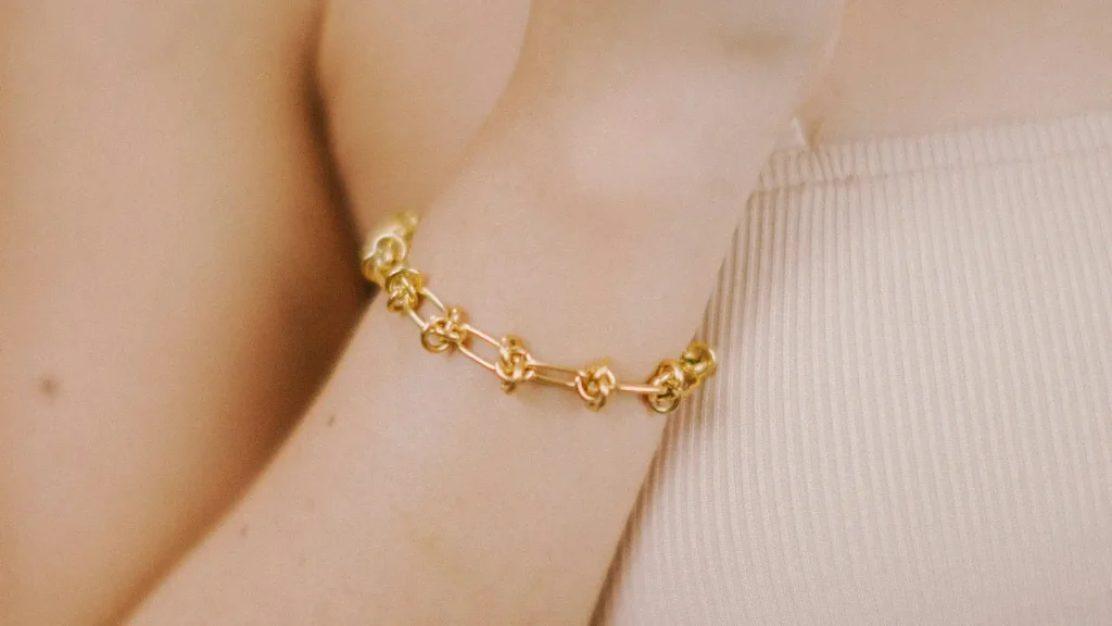 Pulso de pele clara com uma pulseira de ouro de corrente grossa e decorada com detalhes em nós, sobre uma roupa de malha canelada.