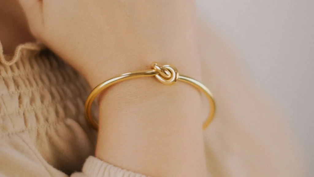 Pulso de pele clara com uma pulseira de ouro rígida e elegante, com um nó decorativo central, sobre um tecido de roupa creme.