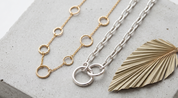 Fotografia de natureza morta sobre base de concreto cinza, exibindo um colar de ouro com elos circulares de textura torcida e uma corrente de prata com elos retangulares e grandes fechos circulares.