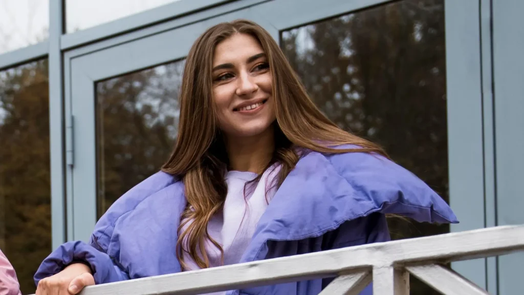 Mulher sorrindo apoiada em uma grade, usando casaco roxo e blusa clara, em ambiente externo, mostrando combinação suave dentro das cores que combinam com roxo.