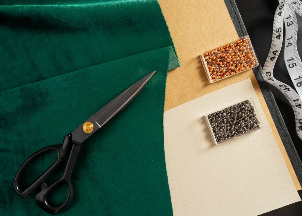 Mesa com tesoura, alfinetes, fita métrica e recortes de tecidos em cores neutras e verde, ilustrando materiais usados com tipos de tecido.