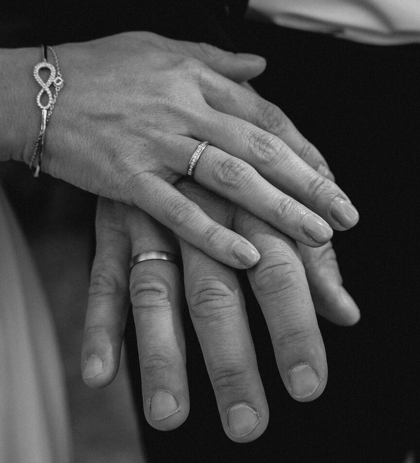 Fotografia em preto e branco focada em close-up nas mãos sobrepostas de um casal. A mão feminina repousa delicadamente sobre a mão masculina. Ela exibe um anel fino cravejado com pequenas pedras e uma pulseira rígida com o símbolo do infinito em destaque. O homem usa uma aliança ou anel de banda lisa e polida.