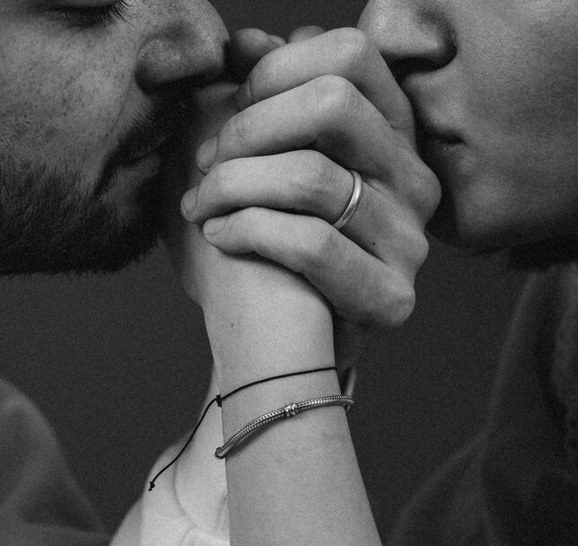 Fotografia expressiva em preto e branco e em plano fechado de um casal com os rostos muito próximos. O homem, com barba curta, encosta os lábios suavemente nos dedos entrelaçados do casal. A mão que está no topo usa um anel liso e largo de prata ou ouro branco. No pulso em evidência, nota-se uma pulseira delicada estilo bracelete de malha de serpente metálica e um cordão fino preto.