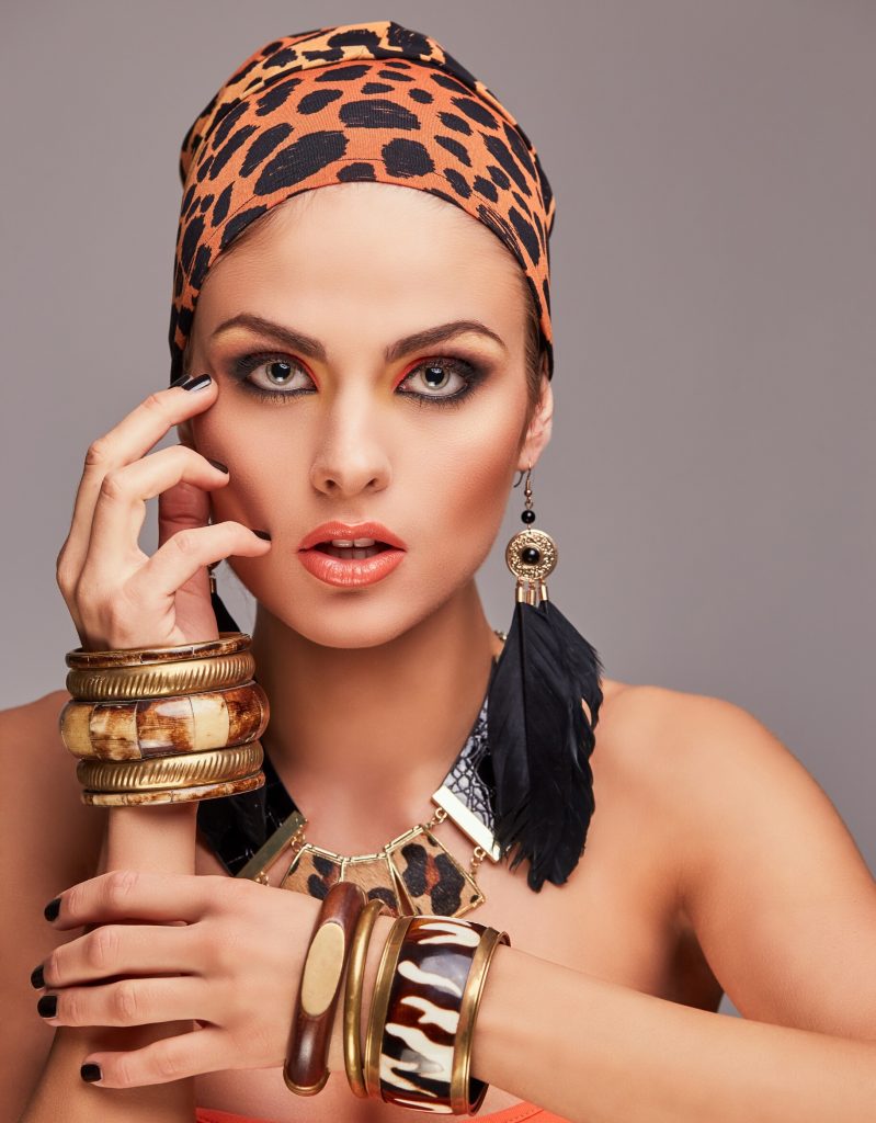 Mulher de pele clara com maquiagem marcante e olhos esfumados, usando um lenço de estampa de onça na cabeça. Ela exibe joias maximalistas: brincos grandes com franjas pretas, colar de elos dourados com detalhes em animal print e múltiplos braceletes largos e empilhados nos dois pulsos, combinando tons de dourado, madeira e estampa de zebra. Fundo cinza neutro.