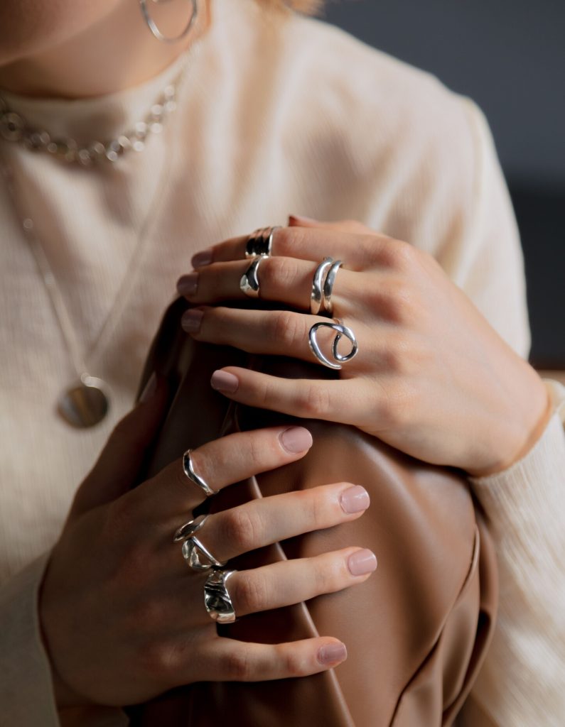 Close de mãos femininas com esmalte nude descansando sobre uma superfície de couro marrom. Ela usa múltiplos anéis grossos de prata com formas orgânicas e fluidas nos dedos, além de uma gargantilha de elos ao fundo.