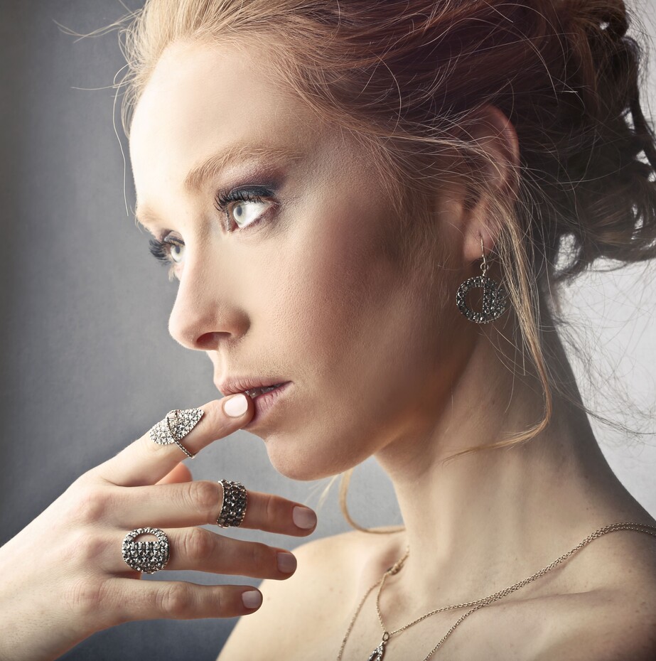 Close de perfil de uma mulher de pele clara e cabelos ruivos presos. Ela usa brincos redondos texturizados e três anéis diferentes com detalhes brilhantes na mão, enquanto toca levemente os lábios. Um colar fino com pingente aparece no colo.