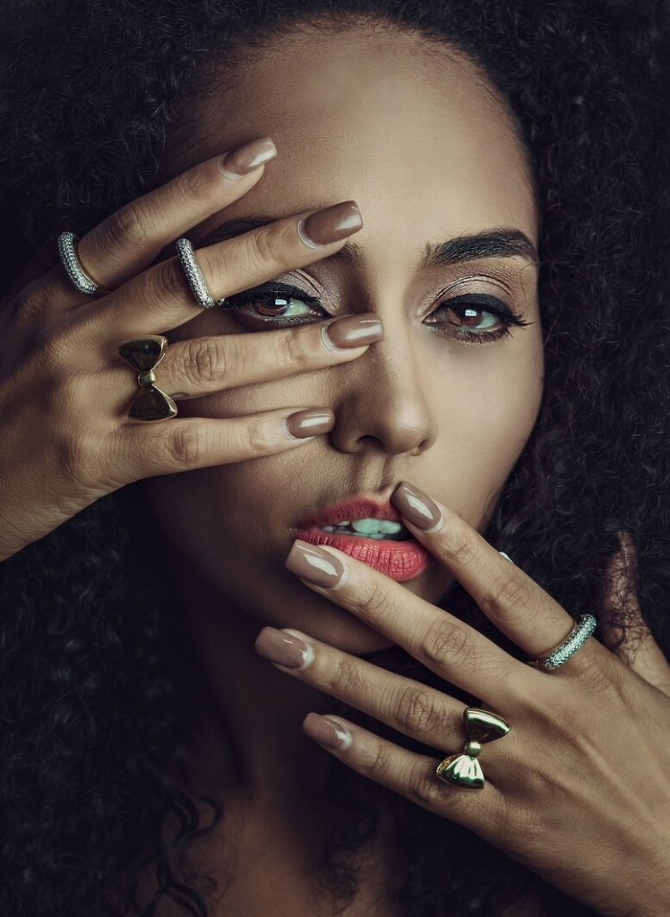 Close do rosto de uma mulher negra de cabelos cacheados volumosos. Suas mãos estão próximas aos olhos e boca, exibindo unhas longas em tom nude e anéis dourados em formato de laço combinados com anéis finos cravejados.