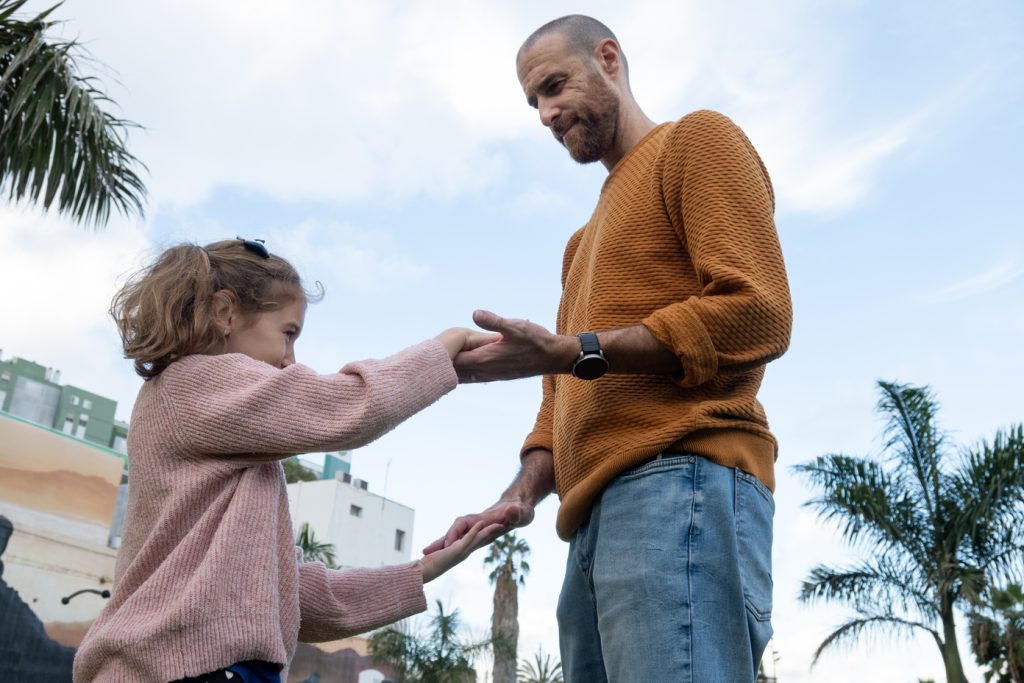 Homem de barba curta e blusa de tricô mostarda visto de um ângulo baixo em um ambiente externo com palmeiras. Ele segura as mãos de uma menina pequena de casaco rosa claro que sorri para ele. O homem usa um relógio de pulseira preta no pulso.