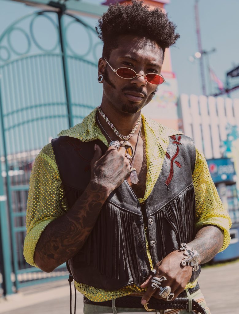 Homem jovem negro com cabelo estilo fade alto posando ao ar livre em frente a um portão de ferro azul. Ele veste um colete de couro preto com franjas sobre uma camisa amarela brilhante com textura de rede. Ele usa óculos de sol ovais de lentes vermelhas, múltiplos anéis grandes de metal nos dedos, braceletes e um colar de corrente grossa com pingente de pedra.