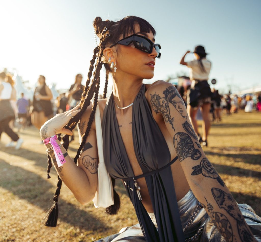 Mulher jovem e tatuada posando agachada em um gramado ensolarado de festival de música com pessoas ao fundo. Ela usa óculos de sol pretos futuristas, um top cinza escuro transpassado com decote profundo e calça metalizada prateada. O cabelo está preso em tranças longas ornamentadas com pequenos anéis metálicos e ela usa brincos pendentes de cruz.