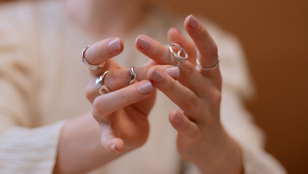 Close das duas mãos de uma mulher com unhas pintadas em tom nude, gesticulando de forma leve. Ela exibe múltiplos anéis de ouro branco com designs diferentes, incluindo formatos lisos, espirais e texturizados, contra um fundo marrom desfocado.