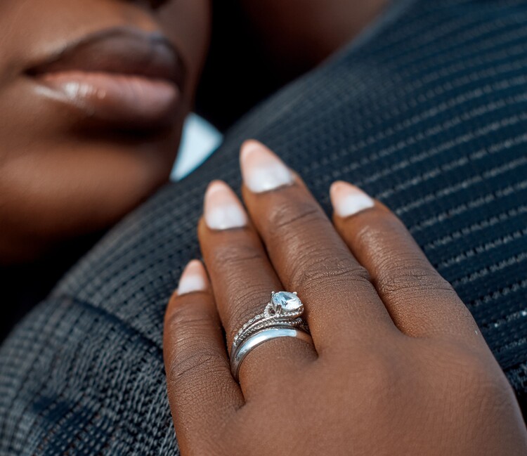Close-up em plano detalhe da mão de uma pessoa negra com unhas estilo francesinha suave, apoiada sobre um ombro com blusa de textura escura. No dedo anelar, destaca-se um anel de noivado de ouro branco cravejado com uma grande pedra de zircônia ou diamante central transparente e lapidada.