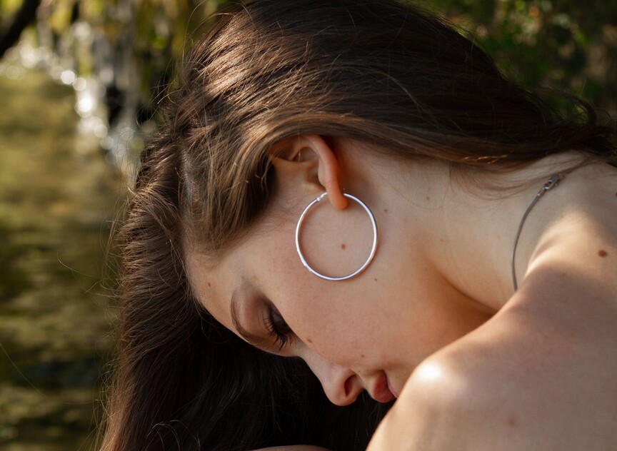 Close de perfil de uma mulher de cabelos castanhos longos ao ar livre, destacando sua orelha com um brinco de argola grande, fino e polido de metal prateado. O fundo mostra uma iluminação suave natural perto d'água.