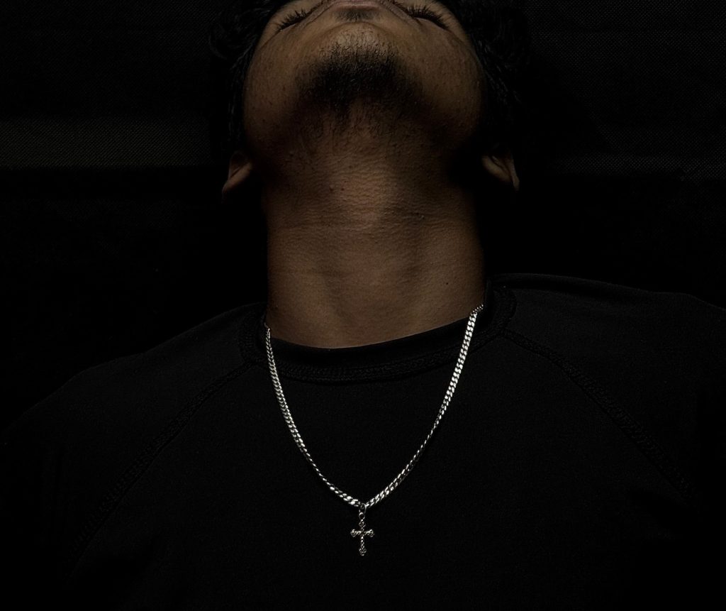 Close-up em plano contra-mergulho do queixo e pescoço de um homem com barba por fazer, olhando para cima. Ele usa uma camisa preta de gola redonda e uma corrente de prata de elos grumet com um pequeno pingente de crucifixo.