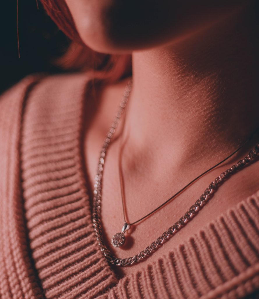 Close-up de uma mulher usando um suéter de tricô rosa empoeirado. Ela usa uma combinação de duas correntes de prata: uma mais curta, tipo cobra, com um pingente redondo de strass, e uma mais longa e grossa, de elos grumet.