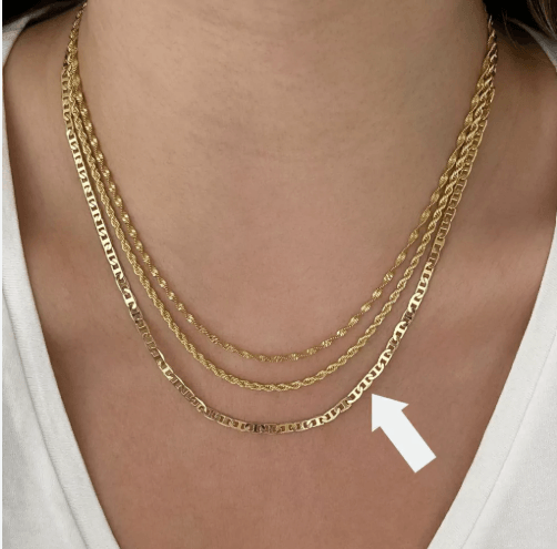 Close-up do colo de uma mulher usando um mix de três colares dourados em camadas, com uma seta branca apontando diretamente para a corrente piastrine de elos planos e achatados na base.