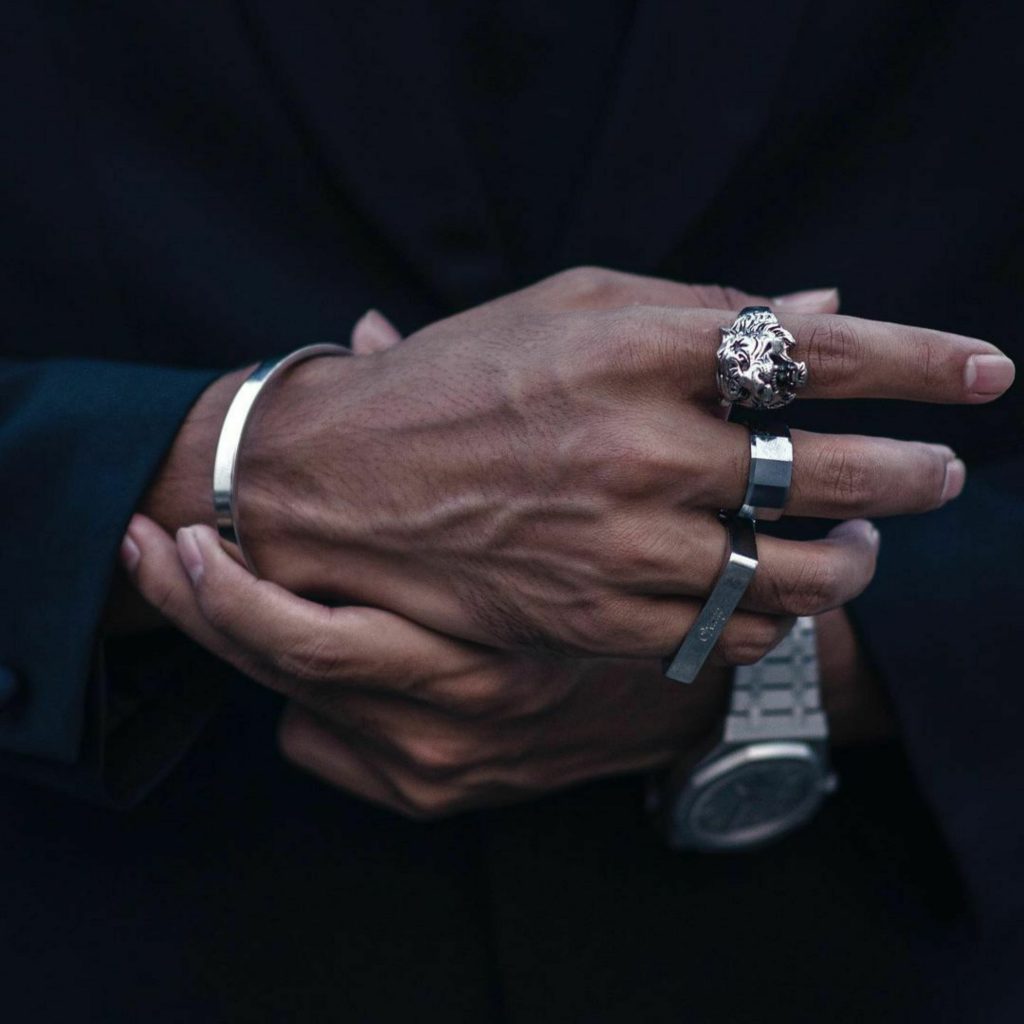 Mão masculina vestindo terno escuro usando mix de anéis de prata, incluindo um anel com formato de tigre, e uma bracelete de prata polida no pulso