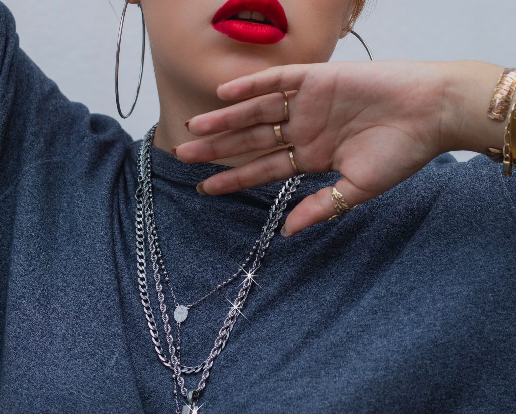 Mulher usando batom vermelho e roupa cinza com mix de correntes de prata em camadas (layering), relicário e grandes brincos de argola.