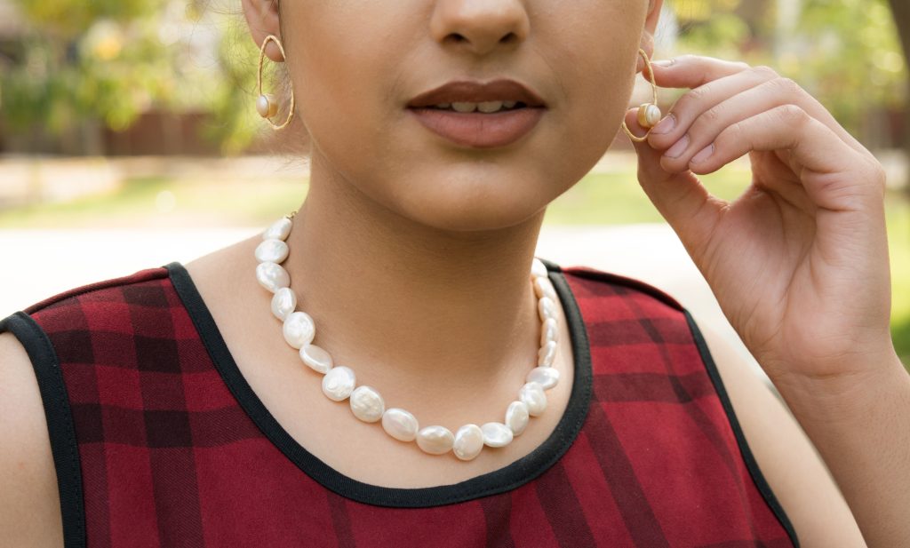 Close-up de uma mulher usando brincos dourados orgânicos com pérolas e um colar curto de pérolas barrocas brancas. Ela veste uma blusa xadrez vermelha e preta, ajustando um dos brincos com a mão.