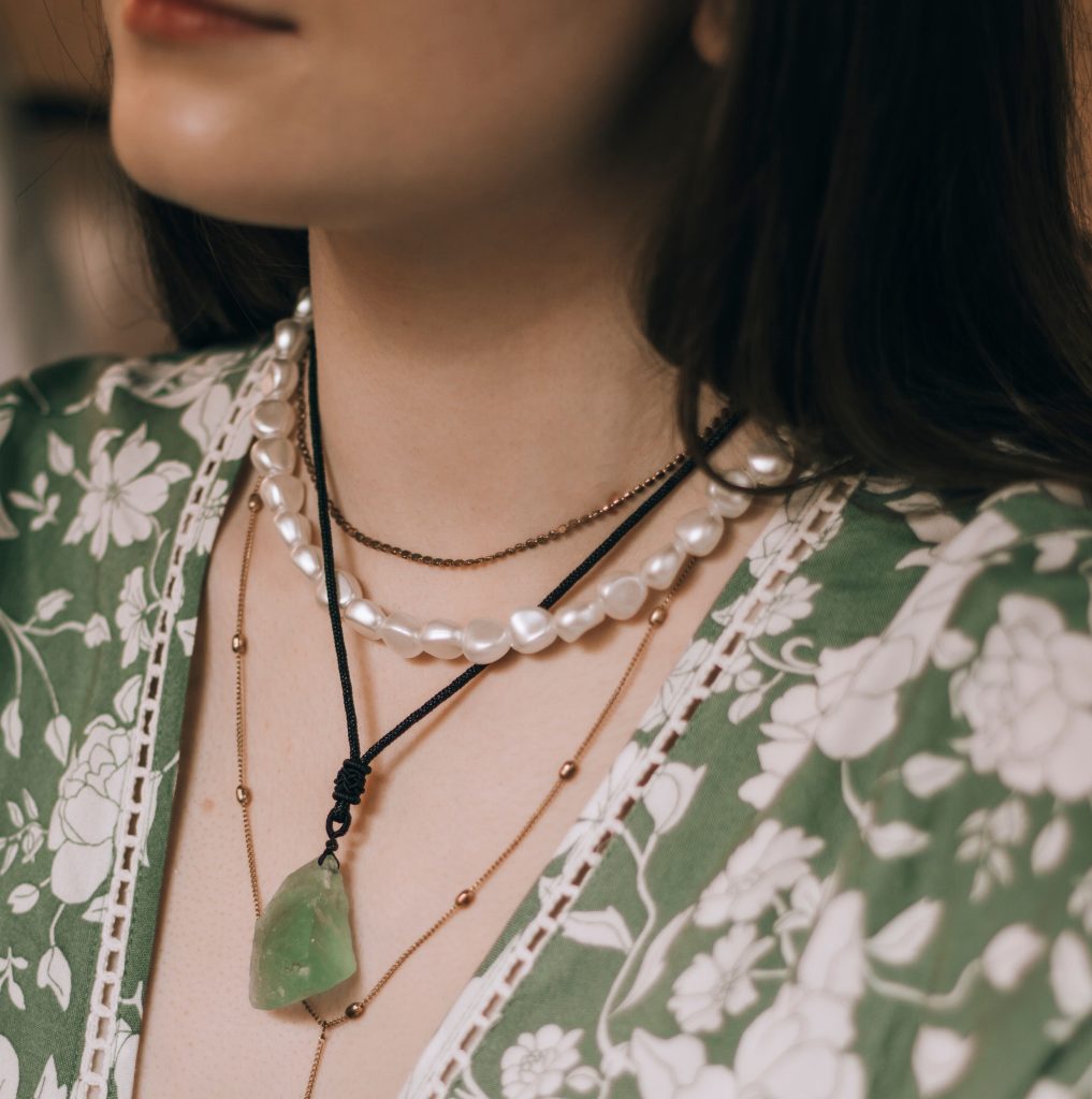 Close-up do colo de uma mulher vestindo uma blusa verde com estampa floral branca. Ela usa um mix de colares em camadas, incluindo uma gargantilha de pérolas barrocas brancas, uma corrente fina escura e um pingente de pedra verde translúcida preso por um cordão.