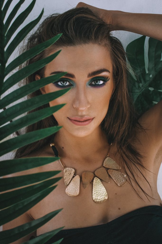 Retrato de uma mulher com maquiagem marcante nos olhos, enquadrada por folhas tropicais verdes, a usar um colar statement dourado com placas metálicas marteladas de formato orgânico e geométrico.