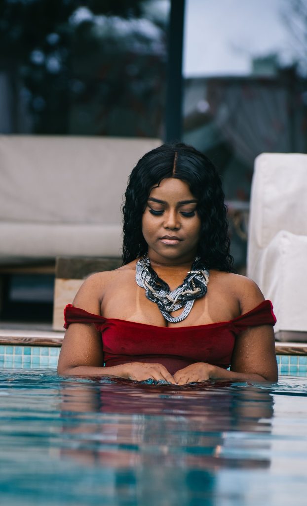 Fotografia de uma mulher negra dentro de uma piscina, a usar um vestido de veludo vermelho ombro a ombro e um maxi colar prateado imponente com múltiplos elos entrançados.