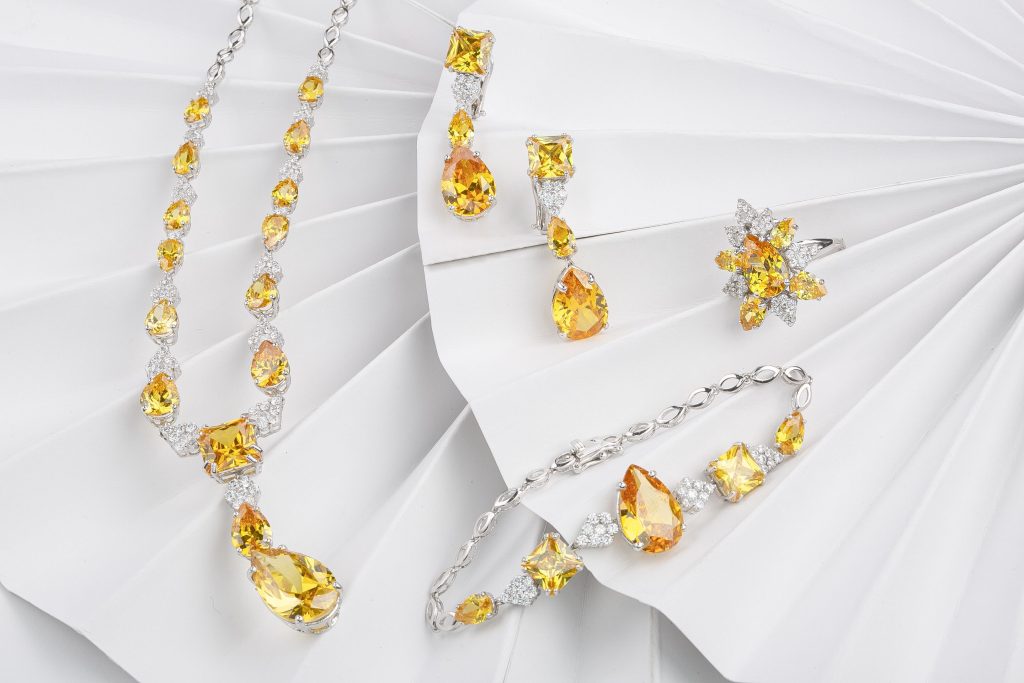 Uma coleção de joias de luxo, incluindo um colar longo, brincos pendentes, um anel e uma pulseira, disposta em uma superfície branca dobrada e texturizada. O conjunto apresenta prata e várias pedras preciosas amarelas facetadas em formas de lágrima e marquise, com acentos claros.