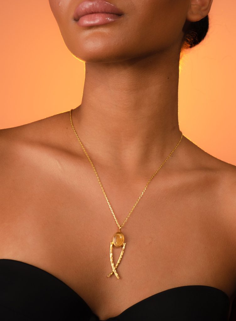 Close-up do busto e pescoço de uma mulher com pele morena, usando um colar dourado. O pingente apresenta uma pedra oval amarela facetada em uma configuração dourada estilizada, contra um fundo laranja quente degradê.